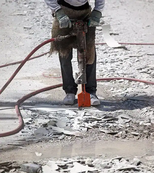 A worker uses a jackhammer to break concrete pavement during construction or renovation work. The scene shows debris and construction tools on a rocky concrete surface.